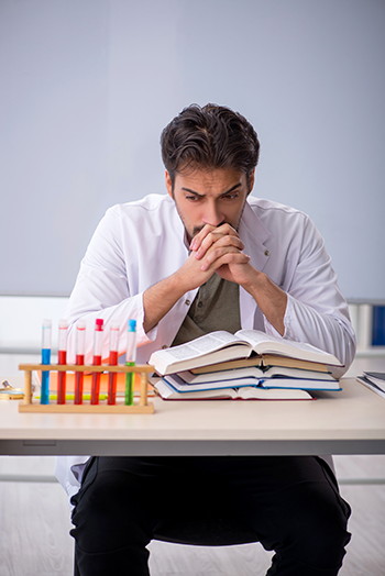 Young male chemist sitting at a table doing research with a pile of books and several beakers filled with various fluids.
